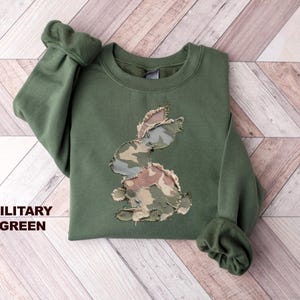 May include: A military green sweatshirt featuring a camouflage bunny design. The sweatshirt has a crew neck and long sleeves with ribbed cuffs. The text "MILITARY GREEN" is displayed on the left side of the image.