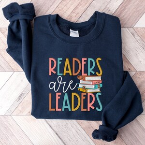 May include: Navy blue sweatshirt with the text "Readers are Leaders" in colorful letters and a stack of books with colorful covers.
