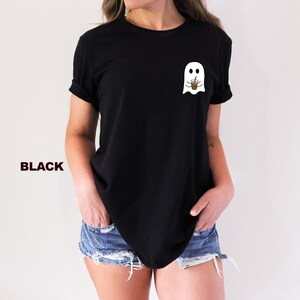 Puede incluir: Una camiseta negra con un fantasma blanco sosteniendo una taza de caf&eacute; en la parte delantera. El texto "BLACK" es visible en la esquina inferior izquierda de la imagen.