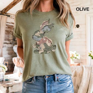 May include: Olive green t-shirt featuring a camouflage bunny design. The bunny is made of patches of fabric in shades of green, brown, and tan. The shirt has rolled-up sleeves and the word "OLIVE" is printed in the upper right corner.