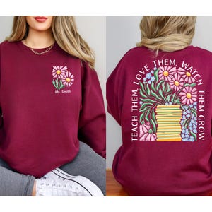 May include: Burgundy sweatshirt with a floral design. The front features a small floral graphic with the name "Ms. Smith." The back displays a larger floral arrangement with the text "Teach them, love them, watch them grow."