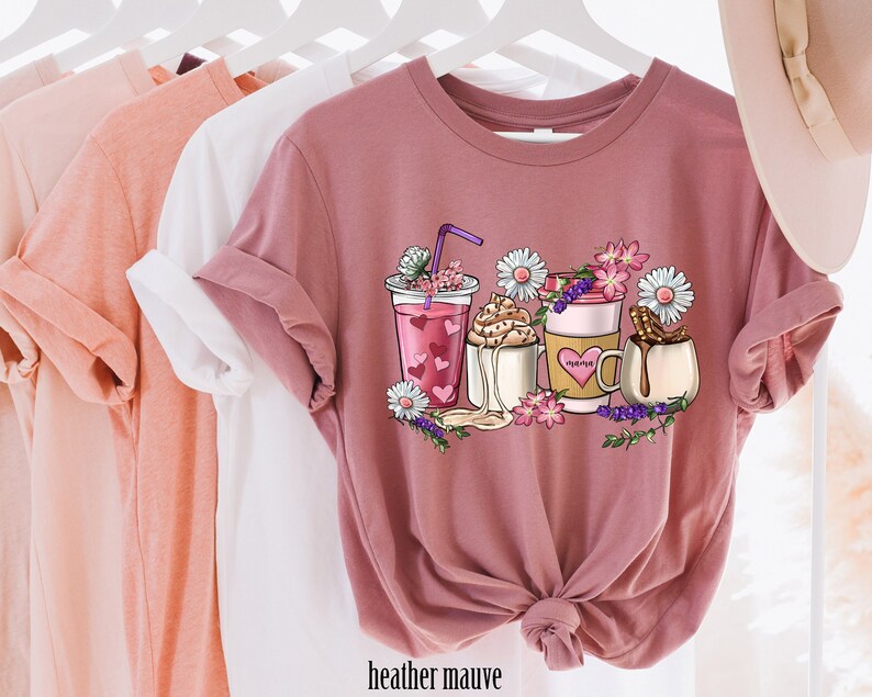 Pode incluir: Uma camiseta de cor malva com um gr&aacute;fico de quatro bebidas de caf&eacute; diferentes com cora&ccedil;&otilde;es, flores e o texto "Happy" em uma das x&iacute;caras.