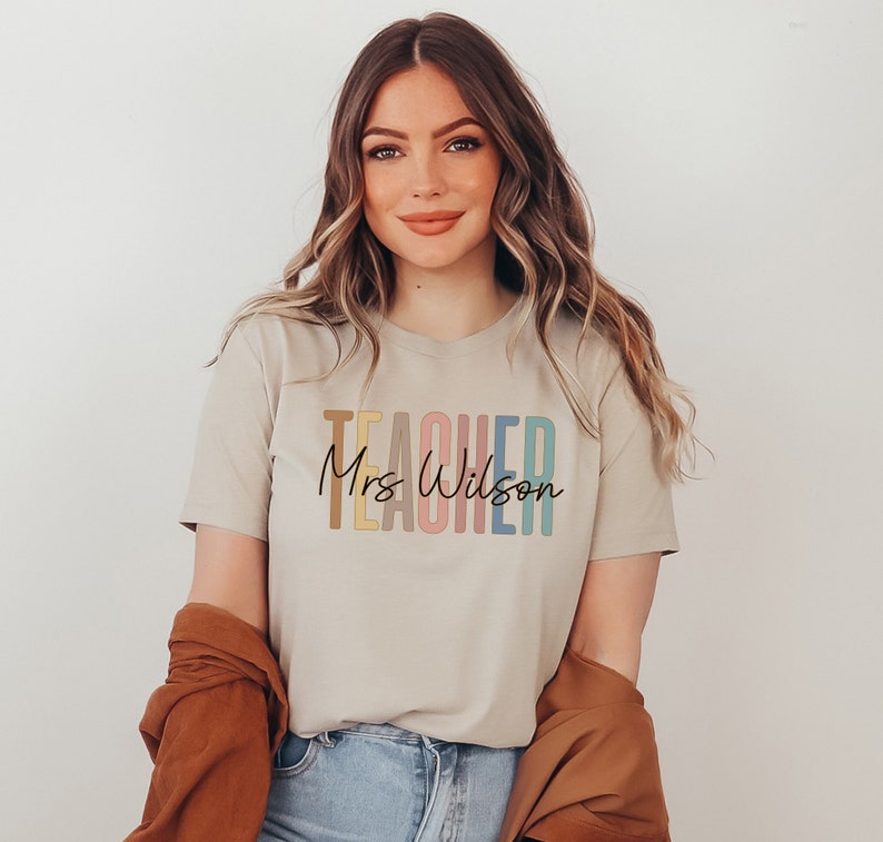 K&ouml;nnte beinhalten: Ein beiges T-Shirt mit dem Wort "TEACHER" in Regenbogenfarben und dem Text "Mrs. Wilson" in schwarzer Schrift.