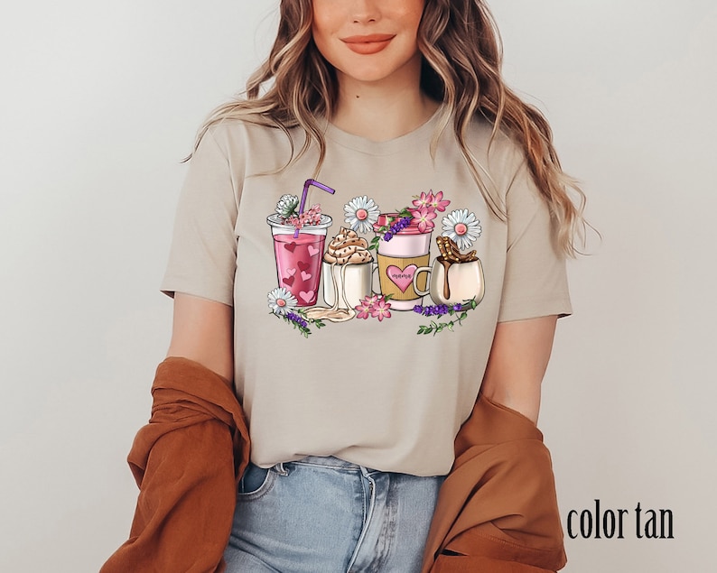 Pode incluir: Camiseta bege com um gr&aacute;fico de quatro bebidas diferentes com flores e um canudo roxo. As bebidas incluem um smoothie rosa, um caf&eacute;, um latte e um chocolate quente.