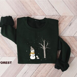 May include: A dark green sweatshirt featuring a white snowman and a small dog in a snowy forest scene. The text "FOREST" is printed on the bottom left corner of the sweatshirt.