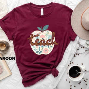 Op de afbeelding: Een maroon t-shirt met een wit appelontwerp met bloemmotieven en het woord "teach" in bruine tekst.