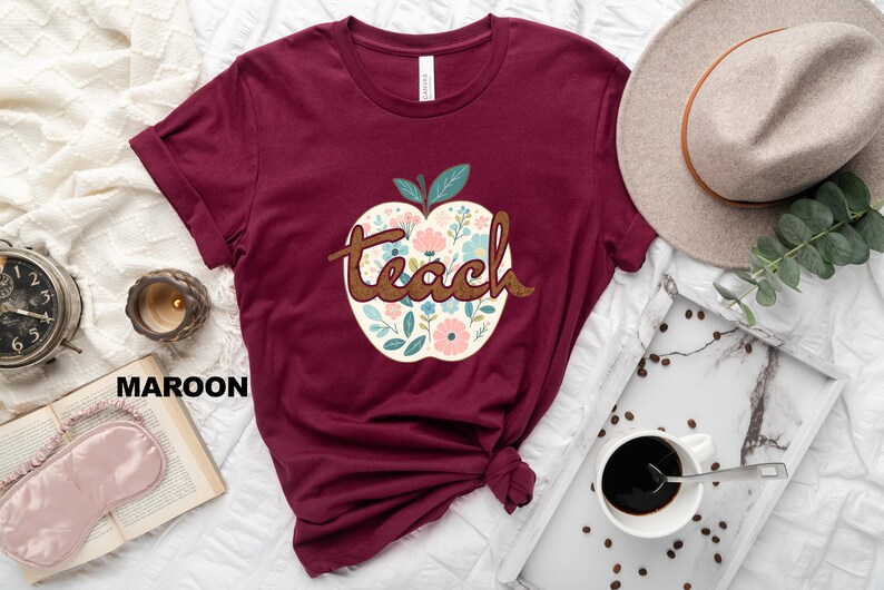Op de afbeelding: Een maroon t-shirt met een wit appelontwerp met bloemmotieven en het woord "teach" in bruine tekst.