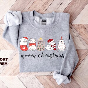 May include: A gray sweatshirt with a Christmas design featuring five cats wearing Santa hats and Christmas lights. The text "merry christmas" is printed below the cats.