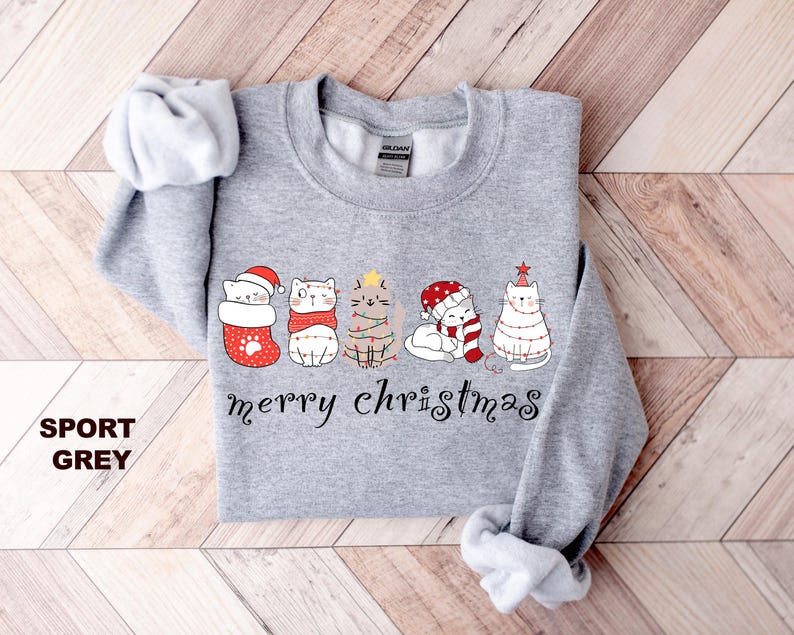 May include: A gray sweatshirt with a Christmas design featuring five cats wearing Santa hats and Christmas lights. The text "merry christmas" is printed below the cats.