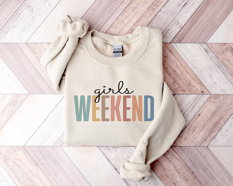 Peut inclure: Un sweat-shirt beige avec le texte "girls weekend" en lettres color&eacute;es. Les lettres sont dans des tons de bleu, jaune, orange et rose.