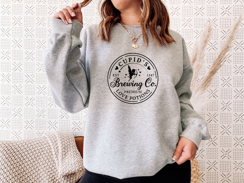 May include: Gray sweatshirt with a black graphic of a circle with the text "Cupid's Brewing Co." and a cupid with an arrow. The text "Est. 1247" and "Premium Love Potions" are also included in the graphic.