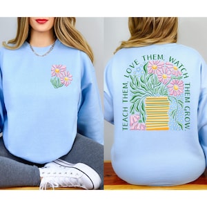 May include: Light blue sweatshirt with a floral design on the front and back. The front features a small cluster of pink and yellow flowers. The back has a larger floral arrangement with the text "Teach them, love them, watch them grow."