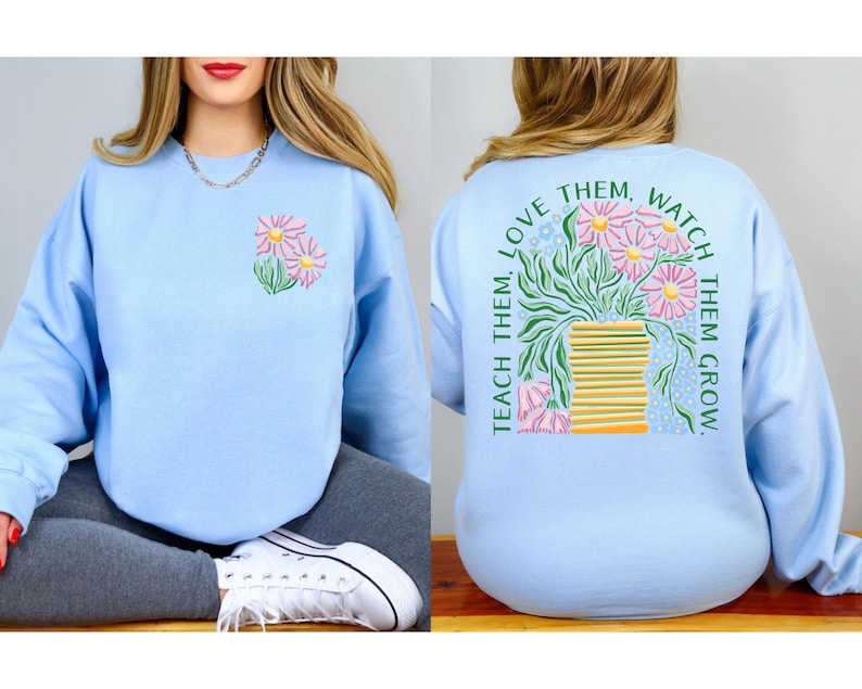May include: Light blue sweatshirt with a floral design on the front and back. The front features a small cluster of pink and yellow flowers. The back has a larger floral arrangement with the text "Teach them, love them, watch them grow."