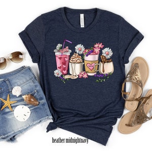 Pode incluir: Uma camiseta azul marinho com um gr&aacute;fico de tr&ecirc;s bebidas diferentes com flores e cora&ccedil;&otilde;es. As bebidas incluem um smoothie rosa, um caf&eacute; com chantilly e um chocolate quente com marshmallows. A camiseta est&aacute; sobre um fundo branco.