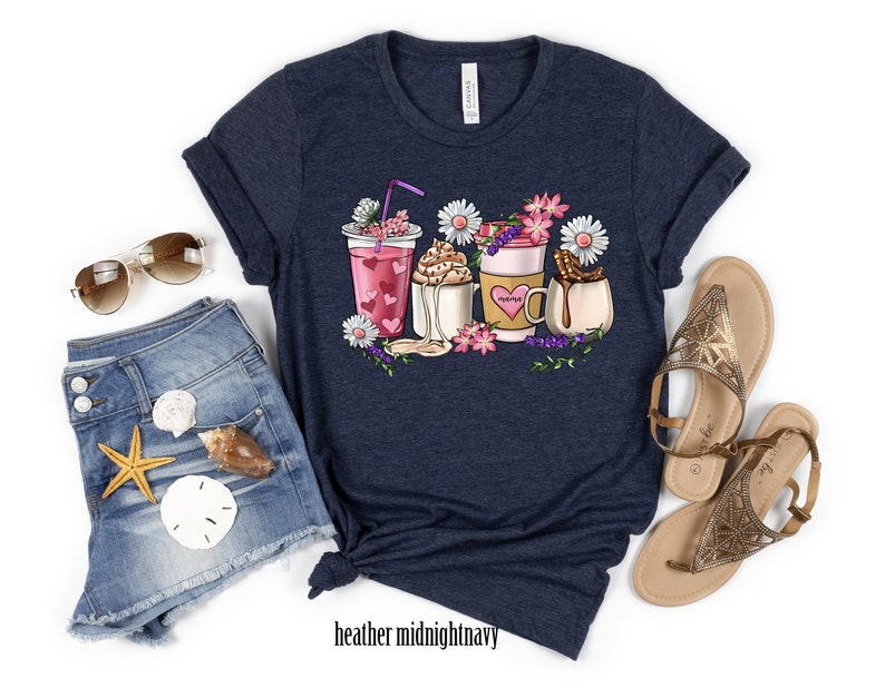Pode incluir: Uma camiseta azul marinho com um gr&aacute;fico de tr&ecirc;s bebidas diferentes com flores e cora&ccedil;&otilde;es. As bebidas incluem um smoothie rosa, um caf&eacute; com chantilly e um chocolate quente com marshmallows. A camiseta est&aacute; sobre um fundo branco.