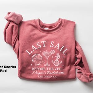 May include: Heather scarlet red sweatshirt with the text "LAST SAIL" above a seashell, cocktail, and anchor graphic. Below the graphic is the text "BEFORE THE VEIL Megan's Bachelorette SAN DIEGO, CA."