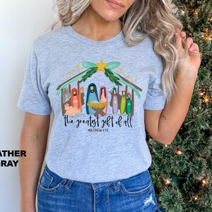 May include: Heather gray t-shirt with a colorful illustration of a nativity scene and the text "The greatest gift of all" with the scripture reference "Matthew 1:21".