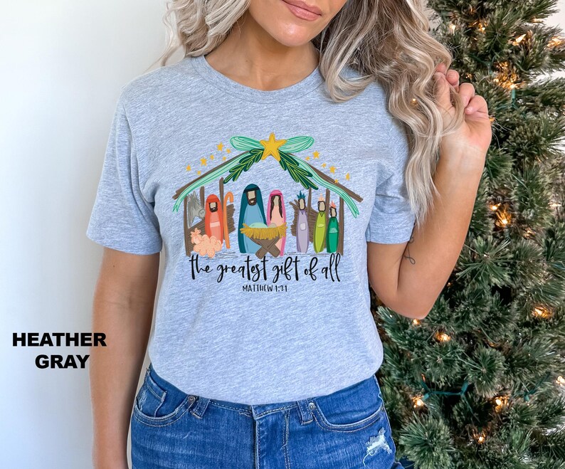 May include: Heather gray t-shirt with a colorful illustration of a nativity scene and the text "The greatest gift of all" with the scripture reference "Matthew 1:21".