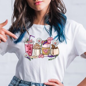 Pode incluir: Uma camiseta branca com um design gr&aacute;fico de quatro bebidas diferentes com flores e um cora&ccedil;&atilde;o em uma das x&iacute;caras. O texto na x&iacute;cara diz "Mam&atilde;e".