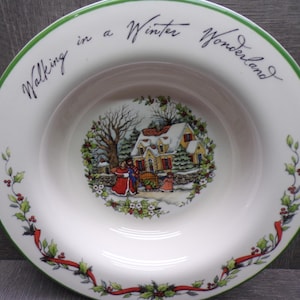 May include: White ceramic plate with a green rim and the words "Walking in a Winter Wonderland". The plate features a winter scene with a house, people, and holly decorations. A red ribbon design adorns the rim.