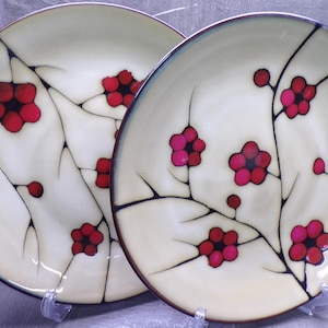May include: Two round ceramic plates featuring a cream-colored base with a hand-painted design of black branches and vibrant red and pink five-petaled flowers. The plates have a dark rim and a smooth, glossy finish across the entire surface.