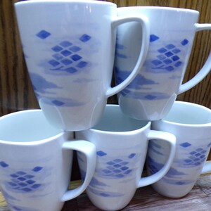May include: Six white ceramic mugs with a blue cloud and diamond design are stacked. Each mug has a large handle and a cylindrical shape. The mugs are stacked on top of each other.