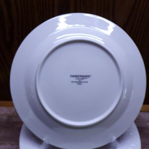 Pu&ograve; includere: Un piatto bianco Farberware Calabar, visto dal basso, con un design circolare. Il piatto ha un bordo largo e un centro leggermente rientrante. Il logo Farberware e le informazioni sul prodotto sono stampati in nero.