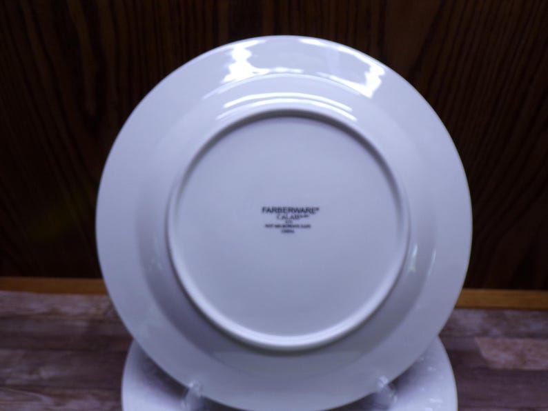 Pu&ograve; includere: Un piatto bianco Farberware Calabar, visto dal basso, con un design circolare. Il piatto ha un bordo largo e un centro leggermente rientrante. Il logo Farberware e le informazioni sul prodotto sono stampati in nero.