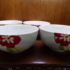 May include: Four white ceramic bowls with a red and yellow floral design. The bowls are round with a wide opening and sit on a dark brown surface. The background is a wooden cabinet.