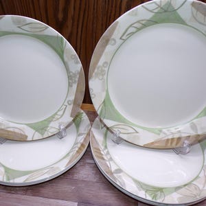 May include: A set of six white ceramic plates with a decorative border featuring green and tan leaf and floral patterns. The plates are displayed on clear plastic stands, showcasing their design.