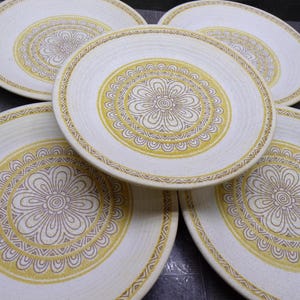 May include: A set of five round ceramic plates featuring a cream-colored base with a central yellow and brown floral mandala pattern. Each plate has a textured surface with concentric circular ridges and a repeating geometric border design.