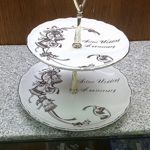 May include: Two-tiered white ceramic cake stand with a gold metal stand. The stand features a silver wedding anniversary design with bells and ribbons. The text "Silver Wedding Anniversary" is printed on both tiers.