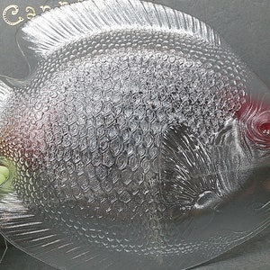 May include: A clear glass fish-shaped serving dish with textured scales and fins. The dish has a red eye and is surrounded by green grapes. The dish is likely used for serving food.