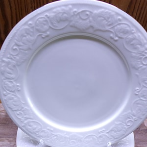 Pu&ograve; includere: Un piatto in ceramica bianca con un motivo decorativo a rilievo di fiori e viti attorno al bordo. Il piatto ha una superficie liscia e lucida e un bordo leggermente rialzato. Il piatto &egrave; probabilmente per servire cibo.