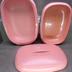 May include: A set of three vintage pink plastic containers. The set includes a large, open container, a perforated insert, and a lid with a slot. The containers are arranged on a gray surface.