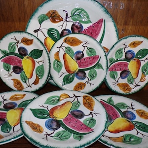 May include: A collection of vintage ceramic plates with a hand-painted fruit design. The plates feature images of pears, plums, and watermelon slices, with green and brown leaves. The plates have a white background with a green border.