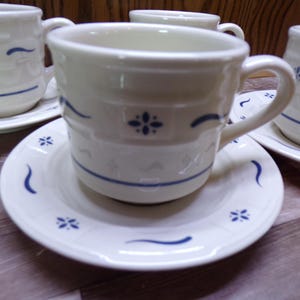 May include: Cream-colored ceramic teacups and saucers with blue floral and wave designs. The cups have handles and are placed on matching saucers. The set is ideal for serving tea or coffee.