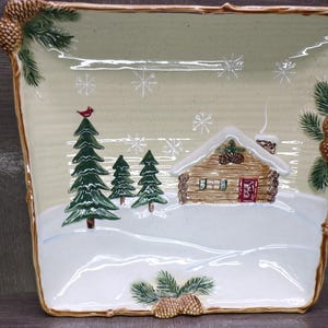 May include: A rectangular ceramic serving platter with a winter scene. The design features a snow-covered landscape with a cabin, pine trees, and snowflakes. The border is decorated with pinecones and greenery.