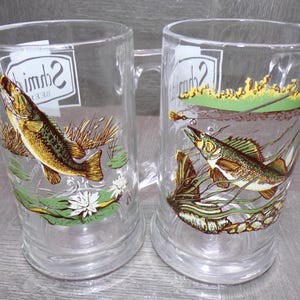 May include: Two clear glass beer mugs with handles. Each mug features a colorful illustration of a fish in a pond setting with aquatic plants. The mugs have the word "Schmidt" printed on them.