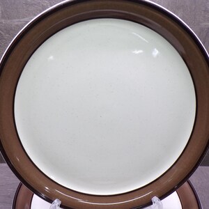 May include: A round ceramic dinner plate featuring a smooth, off-white center with subtle speckles. The plate has a wide, solid brown rim that transitions into a darker brown edge, creating a distinct contrast between the light interior and dark border.
