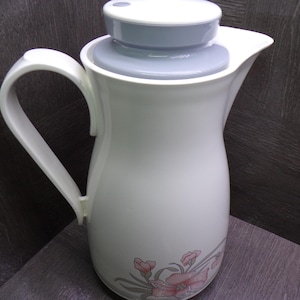 May include: White insulated jug with a curved handle and a pale blue lid. The jug features a floral design in pink and grey near the base. Designed to keep drinks hot or cold, it is ideal for serving.