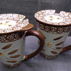 May include: Two ceramic mugs with lids. The mugs are off-white with brown floral and geometric patterns. Each mug has a brown handle and is decorated with brown leaf shapes and yellow flowers. The lids have a cutout for a spoon.