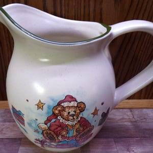 May include: Cream-colored ceramic pitcher with a large handle and a pouring spout. The pitcher features a Christmas-themed illustration of a teddy bear wearing a Santa hat and holding a gift. Green trim accents the rim.