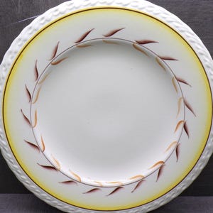 May include: A white ceramic plate with a yellow and brown leaf design. The plate has a textured, white rim and a yellow band around the edge. The leaf design is brown and orange.