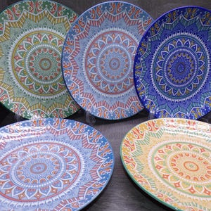 May include: A set of five round ceramic plates featuring intricate, symmetrical mandala patterns in vibrant colors including green, blue, orange, and yellow. Each plate displays a unique radial design with repeating geometric shapes.
