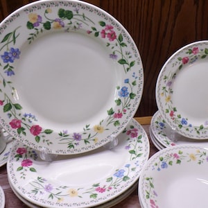 May include: A collection of white ceramic dinnerware featuring a floral design. The plates, bowls, and a teacup are adorned with a border of colorful flowers, including pink, blue, yellow, and purple, set against a backdrop of green leaves.