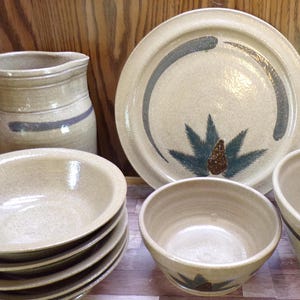 May include: A collection of beige ceramic tableware, including plates, bowls, and a pitcher. The plates feature a blue and gray design with a pinecone and leaf motif. The pitcher and bowls have a simple blue stripe.
