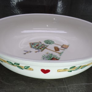May include: White ceramic serving bowl with a decorative rim featuring a green and yellow leaf and branch design. The bowl has a garden scene with a bird, a red heart, and a small house.