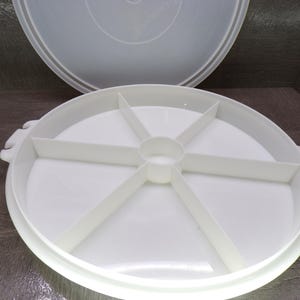 May include: A white plastic serving tray with six compartments and a matching lid. The tray has a round shape with a central hub and dividers. The lid is also white and has a similar round shape. The tray has handles on the sides.