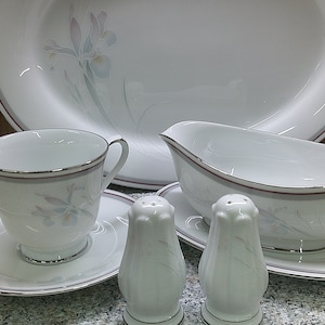 May include: A set of white porcelain dinnerware with a floral design. The set includes a large oval platter, a gravy boat, two teacups and saucers, and two salt and pepper shakers.